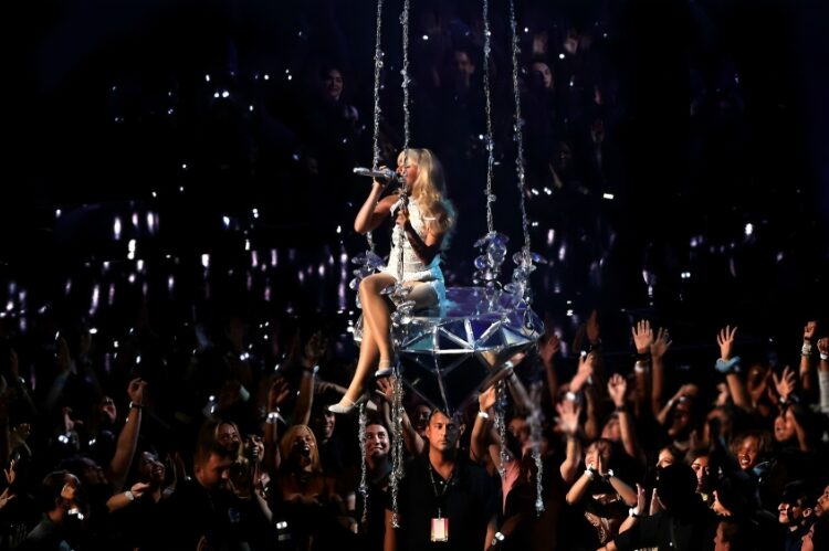 A dramatic concert scene of Sabrina Carpenter in a silver rhinestone corset mini dress with platform boots.