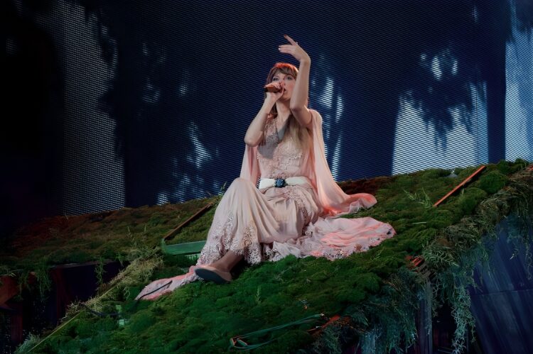 Taylor Swift barefoot in a soft flowy dress, cottagecore style, forest-inspired stage backdrop.