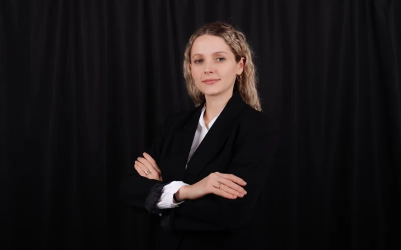 Viktoriia Shykyriava in a black coat, arms crossed, posing confidently against a dark background.
