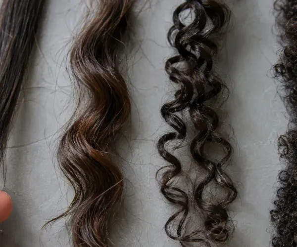 Four natural hair types: straight, wavy, curly, and coily strands on a gray surface.