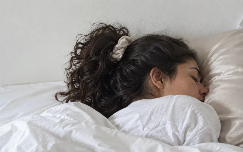 Woman sleeping on her side with long wavy hair tied in a loose pineapple, resting on a satin pillow.