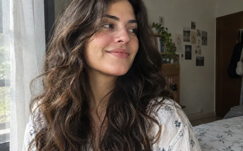 Woman with natural wavy hair smiling by a window in a realistic bedroom.