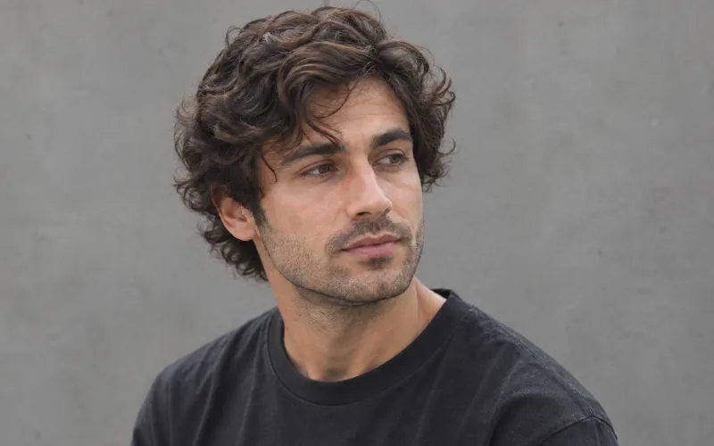 Young man with wavy hair and light stubble against a simple gray background.