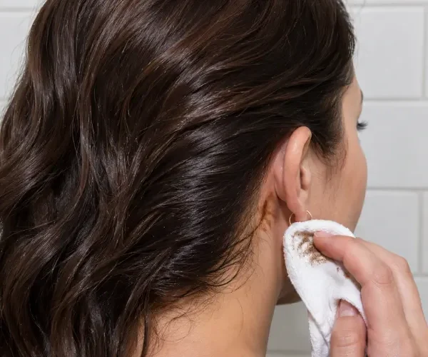 Woman wiping a hair dye stain behind her ear with a white cloth.